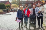 A photograph of a multigenerational family walking down a road.  The father is carrying his toddler daughter and the grandfather pushes a pram as the mother and grandmother talk together.